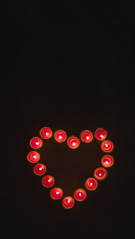 Vertical-Video-Overhead-Shot-Of-Lit-Red-Candles-In-Shape-Of-Romantic-Heart-Being-Blown-Out-On-Black-Background